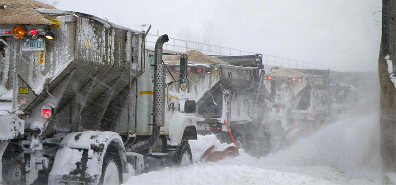 Line of City Plow and Sanding Trucks Heading Up a Street During a Heavy Snowstorm