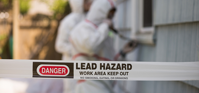 Danger Lead Hazard Tape with Two Workers in Protective Gear Removing Lead Paint From House Behind