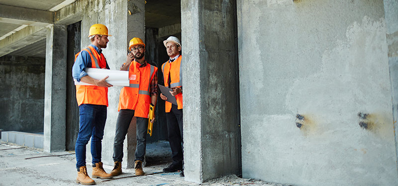 Three People in Safety Vests Looking at a Construction Site and Building Plans