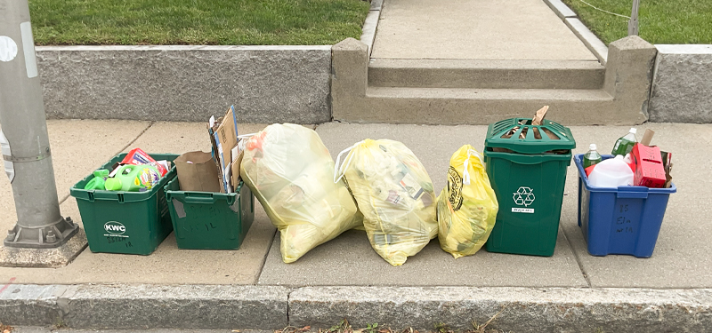 Row of Filled Recycling Bins and Three Yellow City Trash Bags on Street Sidewalk