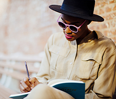 Woman in Hat and Sunglasses Sitting and Writing in a Notebook