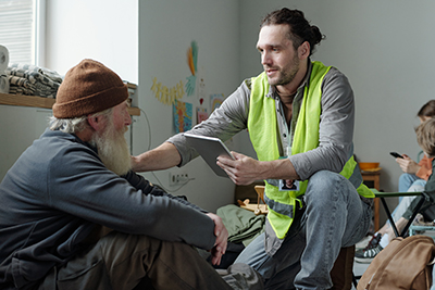 Young Male Volunteer Touching Shoulder of Senior Man Sitting at Temporary Shelter
