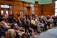 Group of Attendees Sitting in Chairs and Standing in Back Watching State of the City Address at City Hall
