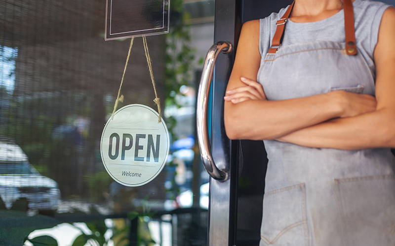 Woman in Gray Apron Standing at Door of Business with Open Sign