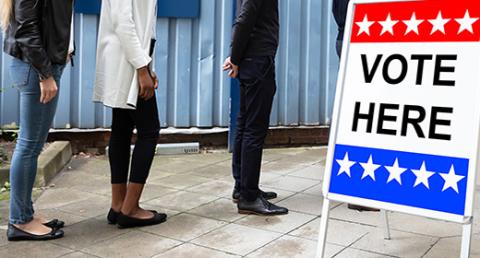 People Waiting in Line next to a Red, White and Blue Vote Here Sign
